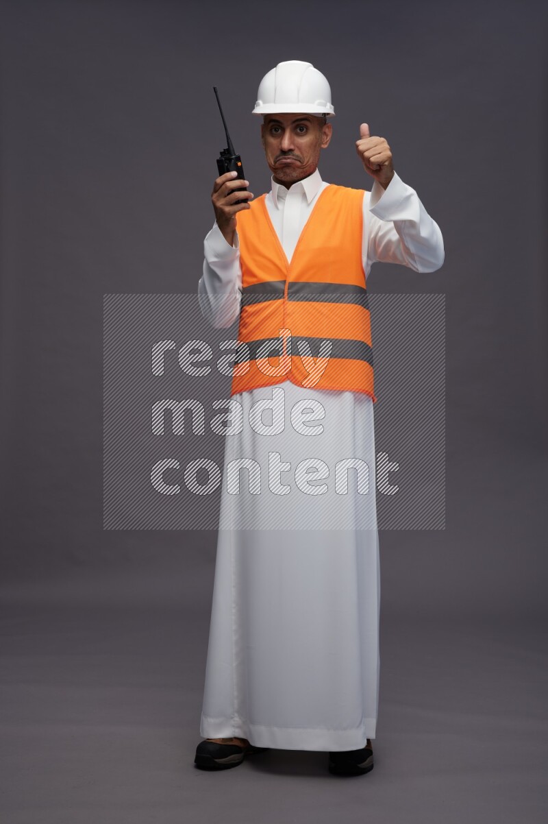 Saudi man wearing thob with engineer vest standing holding walkie-talkie on gray background