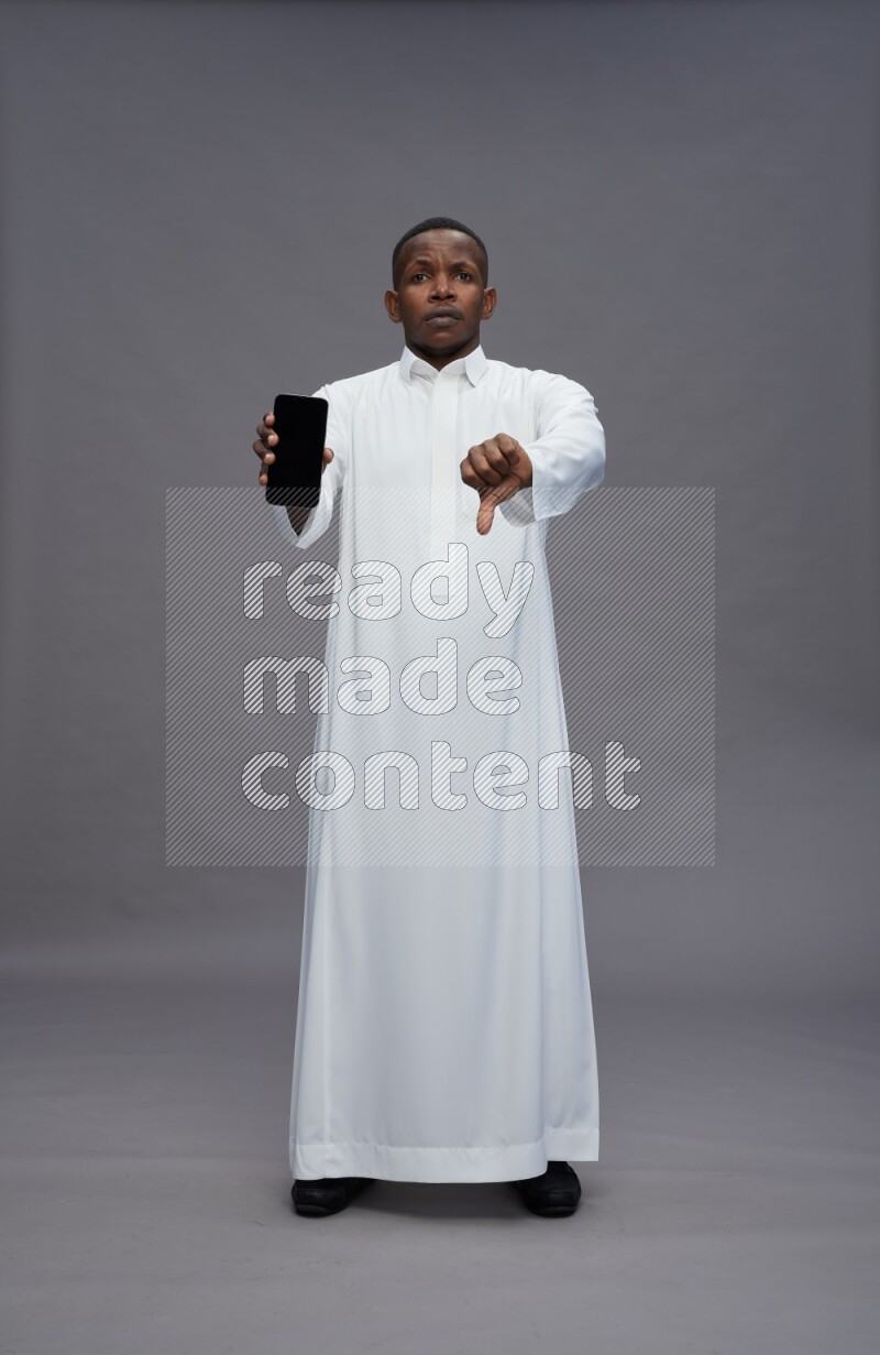 Saudi man wearing thob standing showing phone to camera on gray background