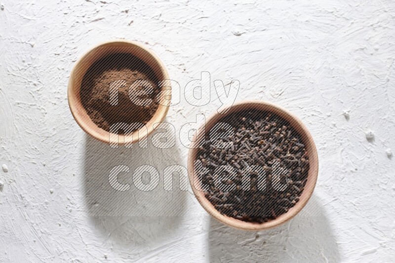 2 wooden bowls full of cloves powder and whole cloves on a textured white flooring