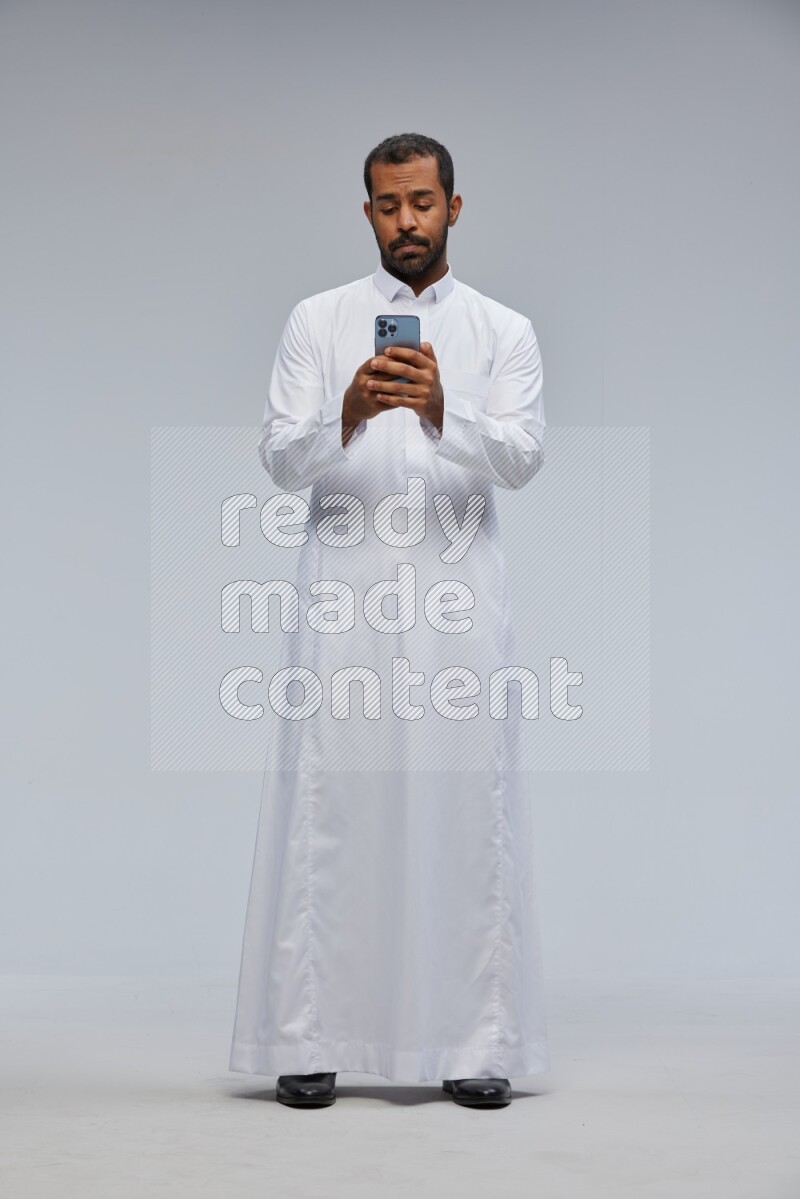 Saudi man wearing Thob standing texting on phone on Gray background