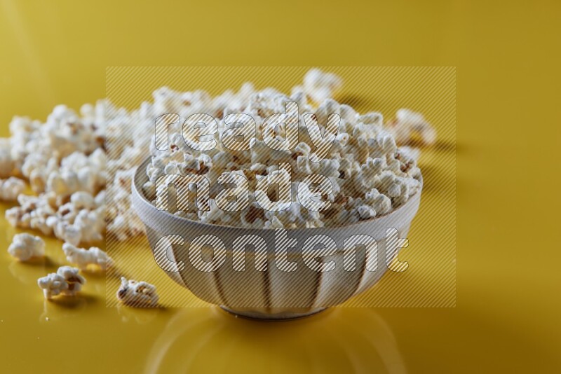 A white pottery bowl full of popcorn with popcorn beside it on a yellow background in different angles