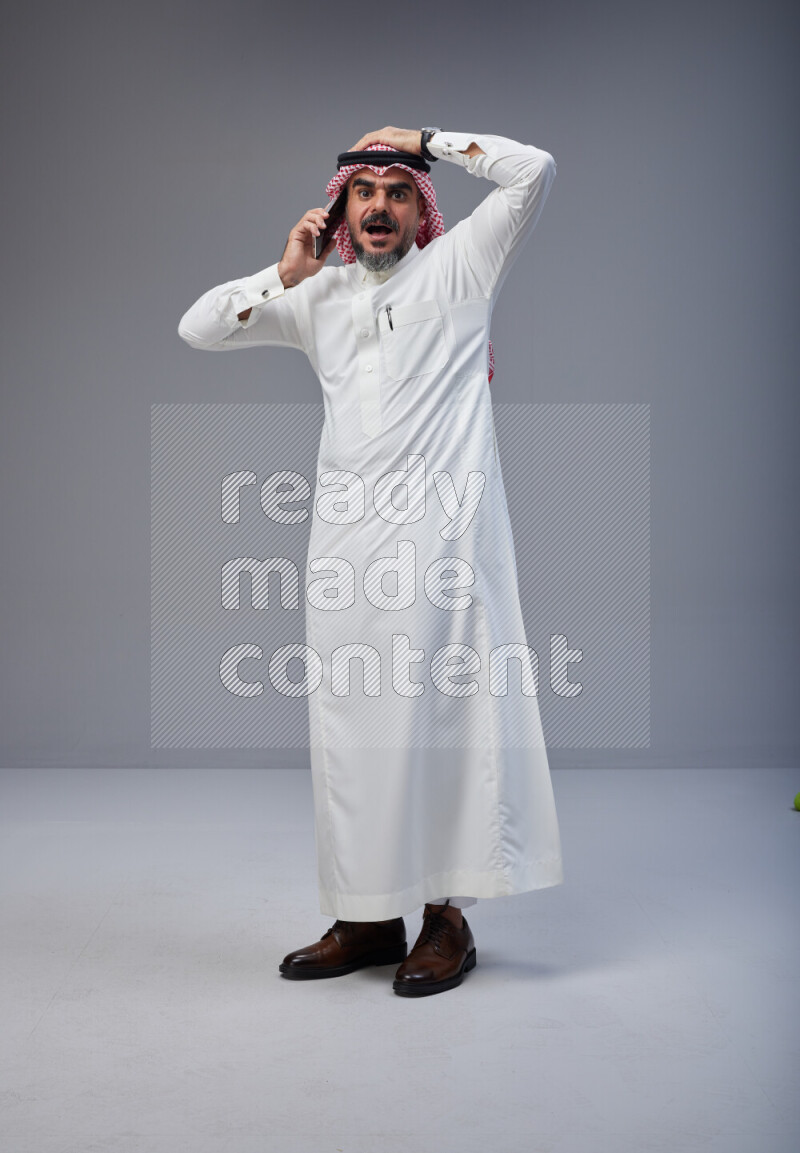 Saudi man Wearing Thob and red Shomag standing talking on phone on Gray background