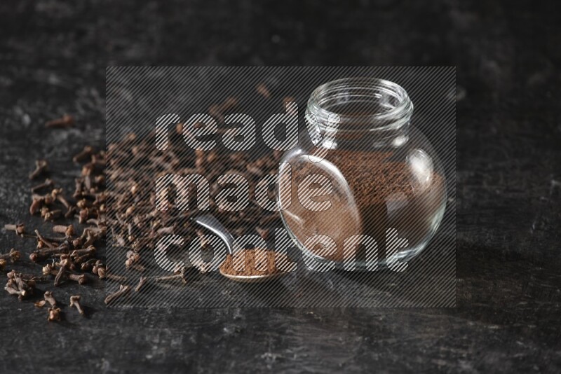 A glass spice jar and a metal spoon full of cloves powder with cloves spread on textured black flooring