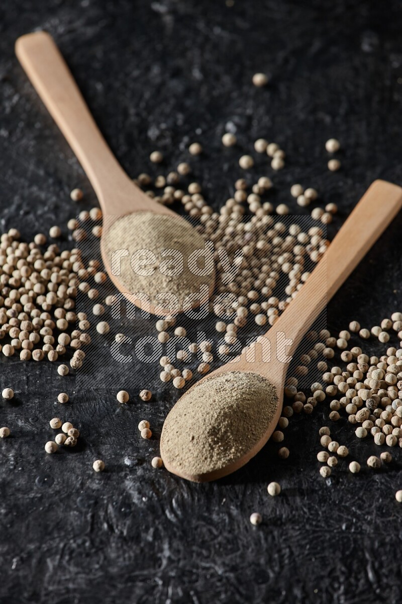 2 wooden spoons full of white pepper powder with white pepper beads on textured black flooring