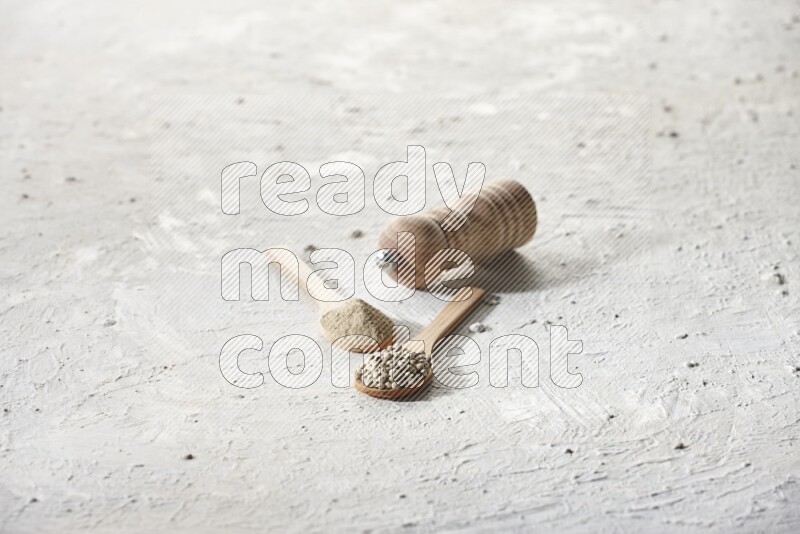 2 wooden spoons one full of white pepper powder and the other with pepper beads and a wooden pepper grinder on textured white flooring
