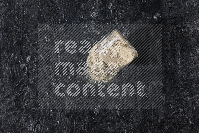 A flipped glass jar full of white pepper powder with spilled powder on textured black flooring