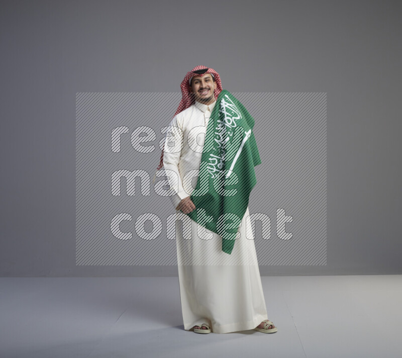 A saudi man standing wearing thob and red shomag holding big saudi flag on gray background