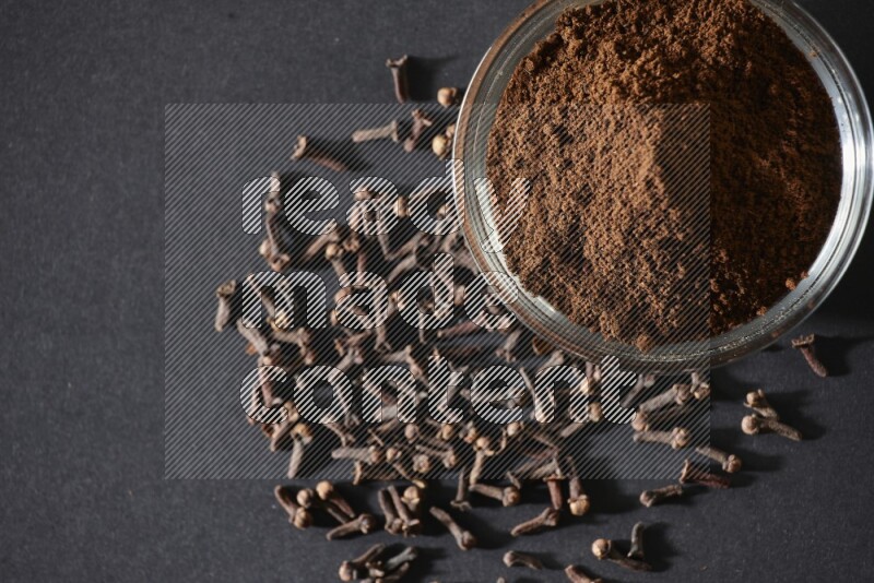 وعاء زجاجي ممتلئ ببودرة القرنفل مع حبوب القرنفل المتناثرة على أرضية سوداء