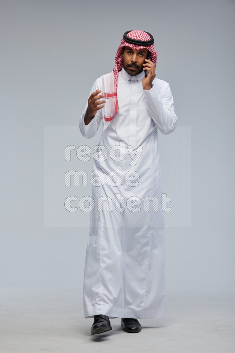Saudi man Wearing Thob and shomag standing talking on phone on Gray background