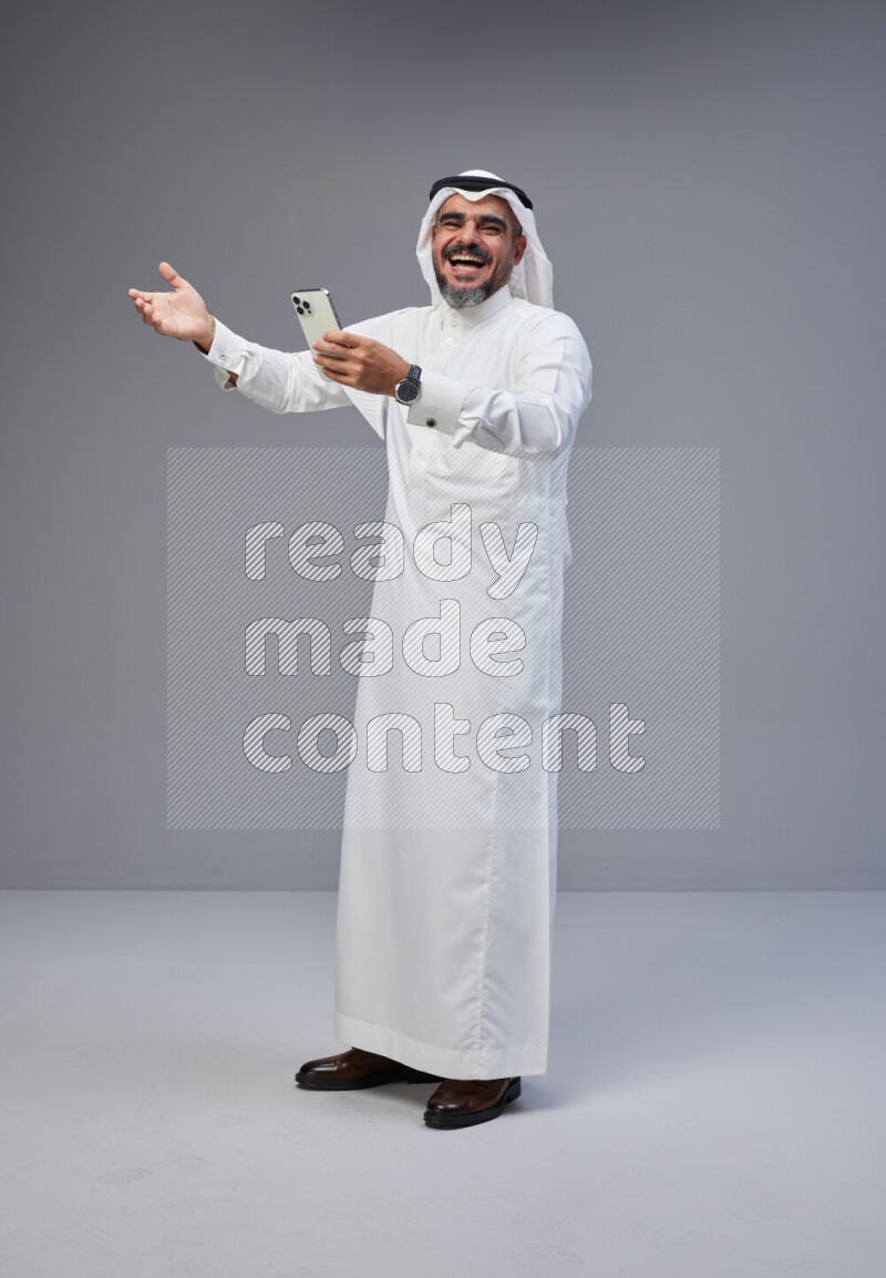 Saudi man Wearing Thob and white Shomag standing texting on phone on Gray background
