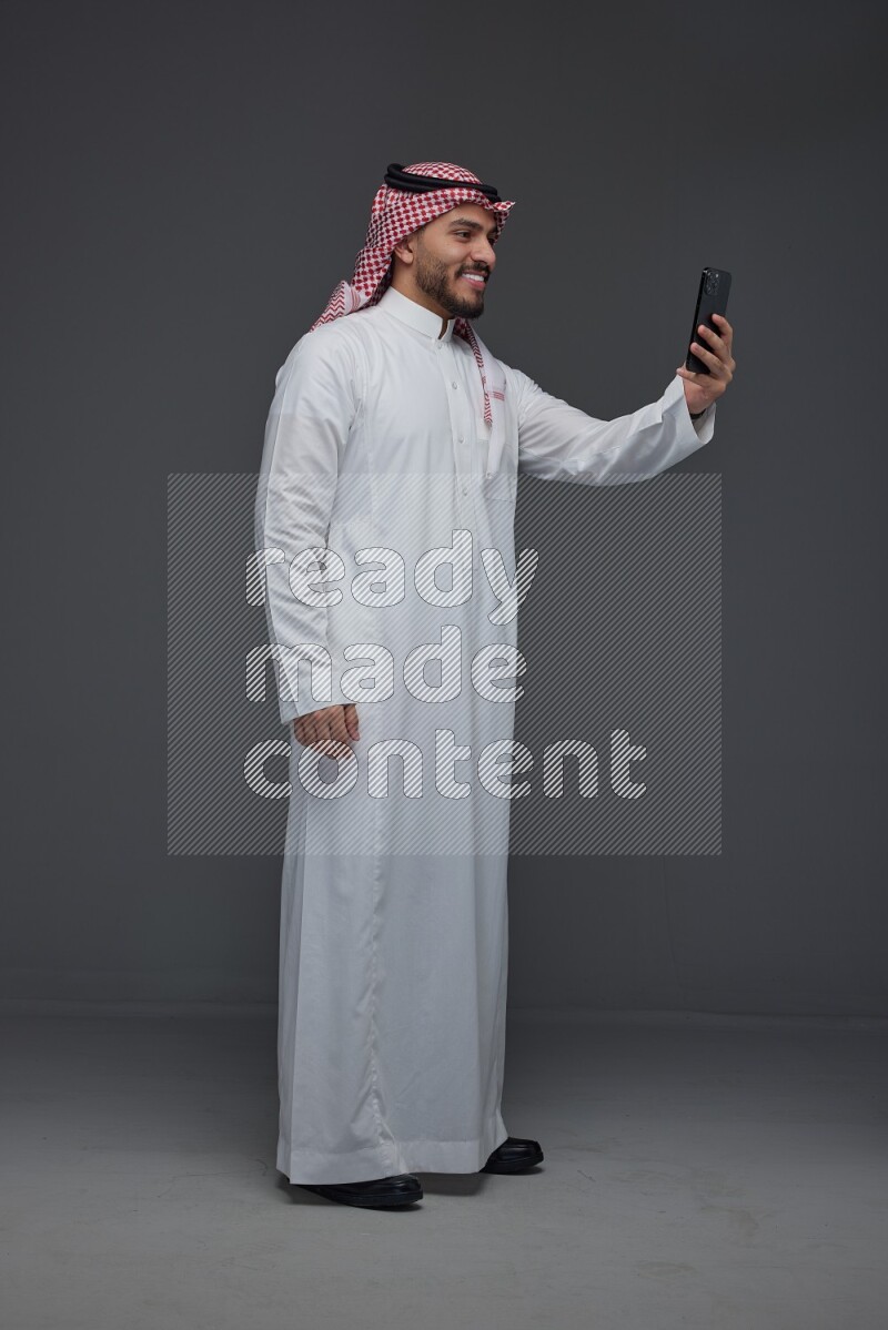 A Saudi man wearing Thobe and Shamgh taking selfie with his phone eye level on a gray background