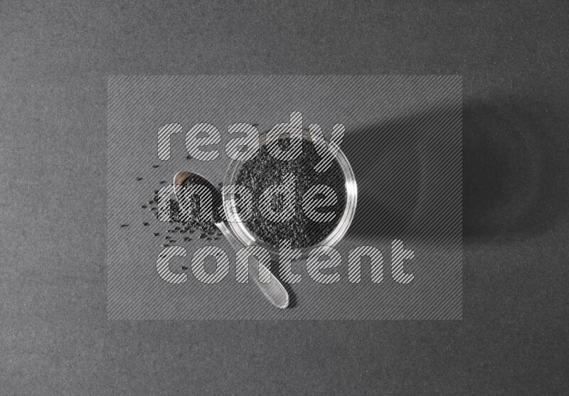 A glass bowl full of black seeds with a metal spoon full of the seeds on a black flooring