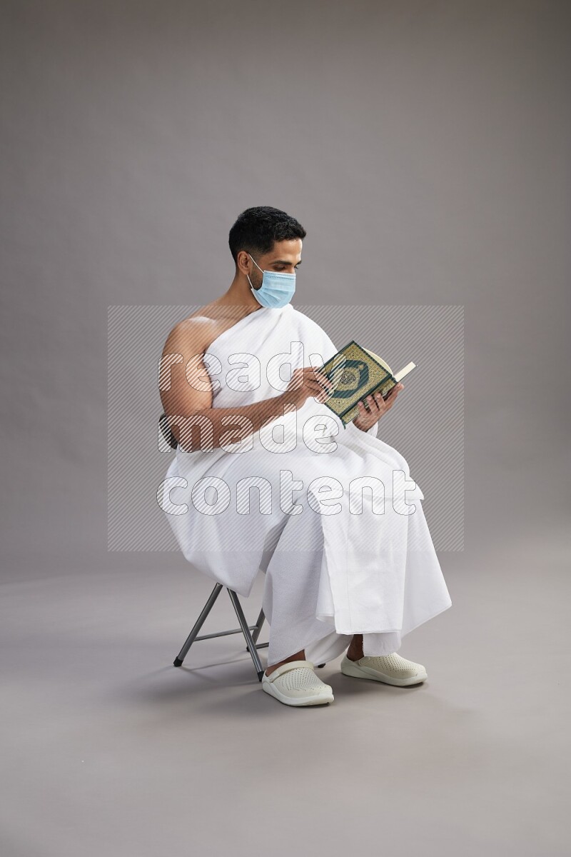 A man wearing Ehram with face mask sitting on chair reading quran on gray background
