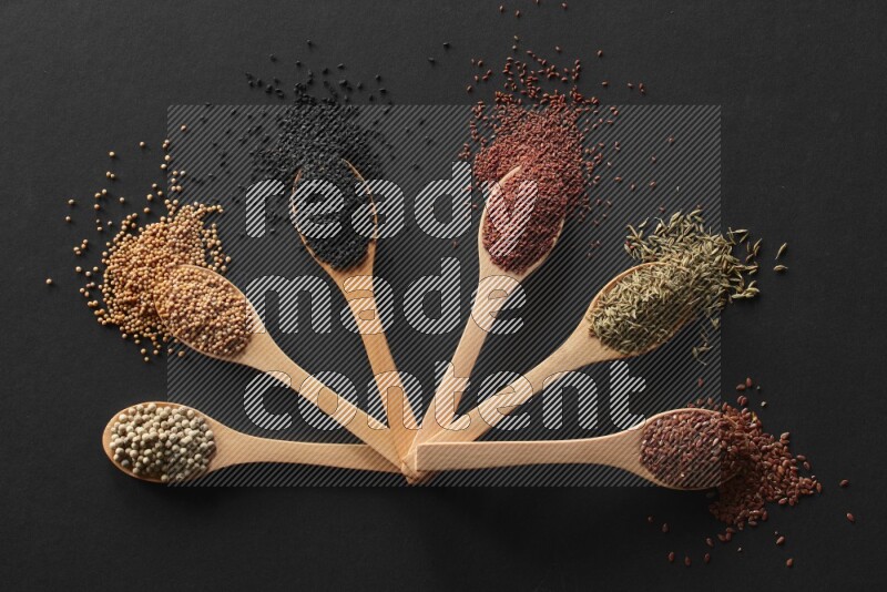 wooden spoons filled with white peppers, mustard seeds, black seeds, garden cress, cumin and flax on black flooring