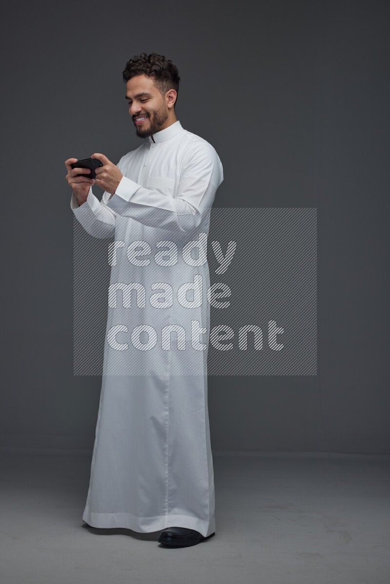 A Saudi man wearing Thobe and playing using his phone horizontally playing video games while standing and making different poses eye level on a gray background