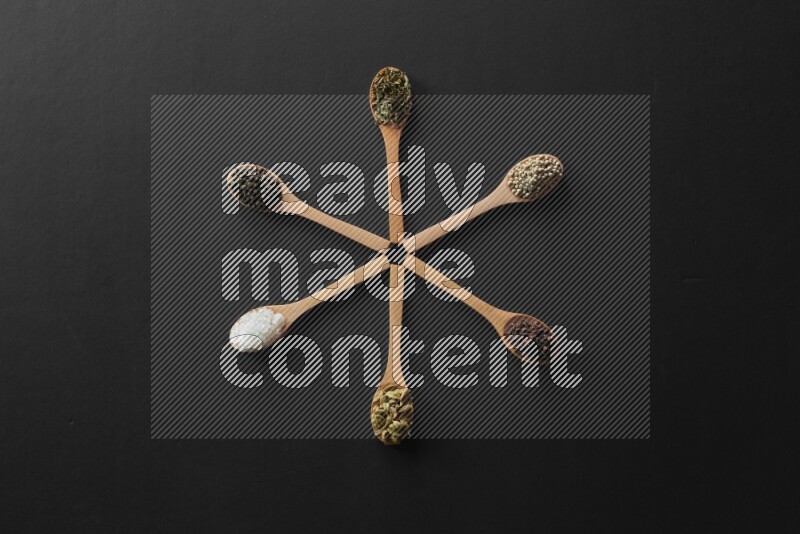 wooden spoons filled with white peppers, cloves, cardamom, salt, black peppers and basil on black flooring