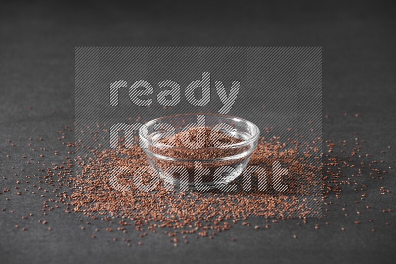 A glass bowl full of garden cress seeds surrounded by seeds on a black flooring