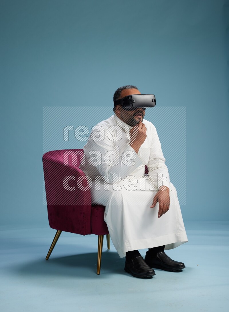 Saudi Man without shimag sitting on chair  gaming with VR on blue background