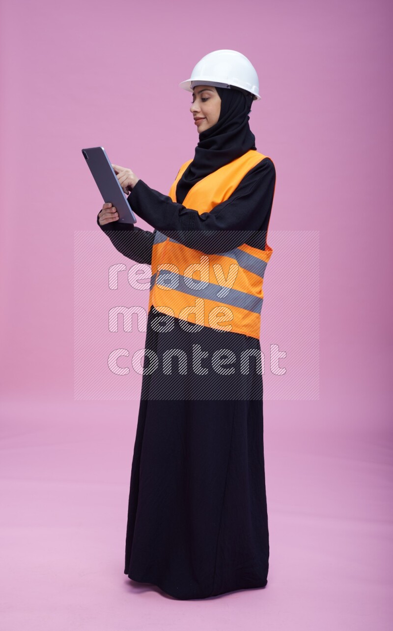 Saudi woman wearing Abaya with engineer vest and helmet standing working on tablet on pink background