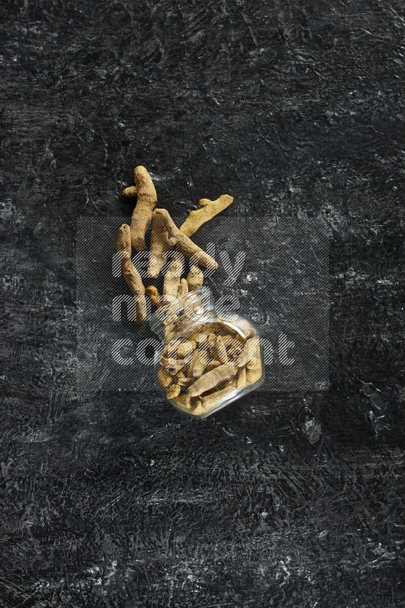 A flipped glass spice jar full of dried turmeric whole fingers and fingers fell out of it on textured black flooring