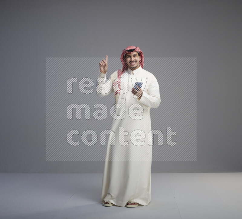 A  Saudi man standing wearing thob and red shomag texting on phone on gray background