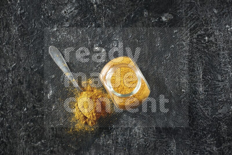 A glass spice jar and metal spoon full of turmeric powder on textured black flooring