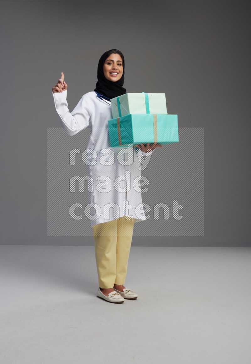 Saudi woman wearing lab coat with stethoscope standing holding gift box on Gray background