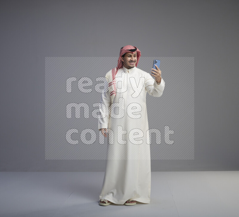 A Saudi man standing wearing thob and red shomag taking selfie on gray background