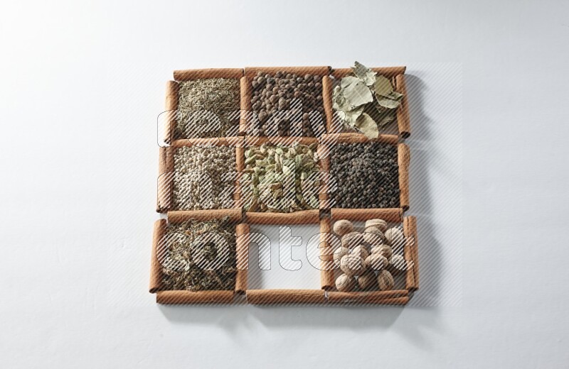 9 مربعات من أعواد القرفة ممتلئة بالهيل في المنتصف محاطة بجوزة الطيب والملح والريحان المجفف والفلفل الأبيض والكمون وبهار الفلفل الحلو وأوراق الغار والفلفل الأسود على أرضية بيضاء