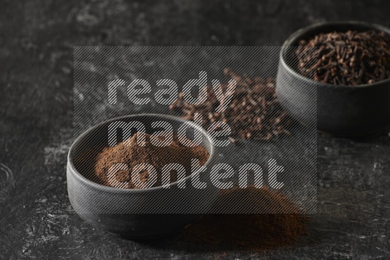 2 Black pottery bowls full of cloves and the other full of cloves powder on textured black flooring