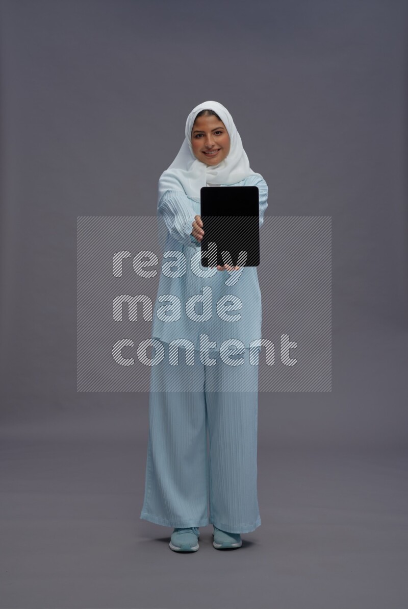Saudi woman wearing hijab clothes standing showing tablet to camera on gray background