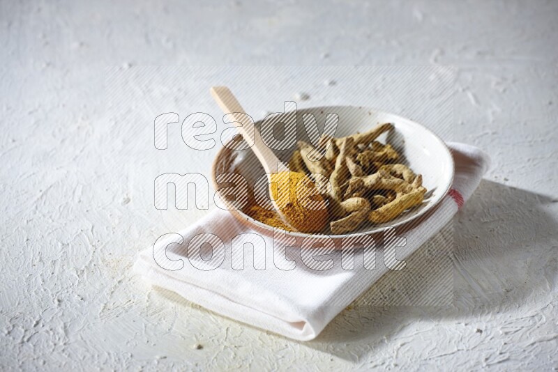 A plate filled with dried turmeric whole fingers and a wooden spoon full of turmeric powder on a textured white flooring
