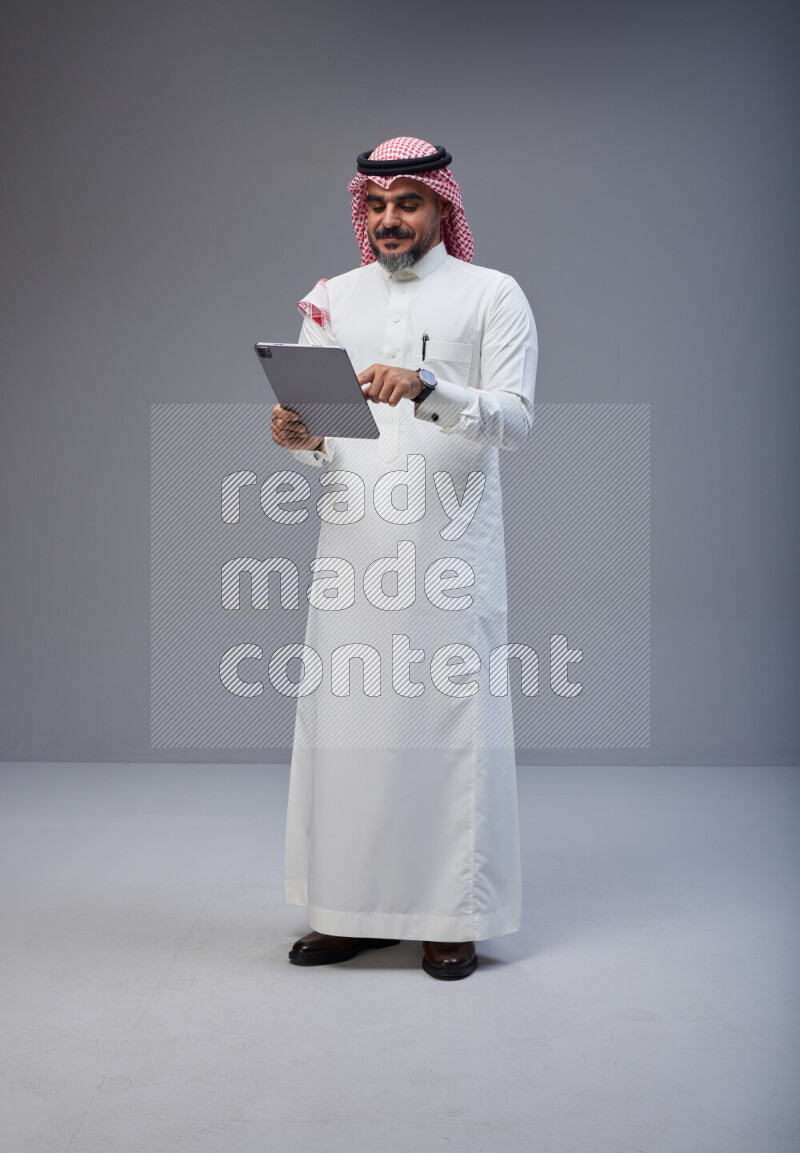 Saudi man Wearing Thob and red Shomag standing working on tablet on Gray background