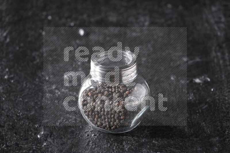 A glass spice jar full of black pepper beads on textured black flooring