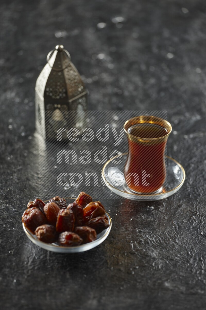 A silver lantern with different drinks, dates, nuts, prayer beads and quran on textured black background