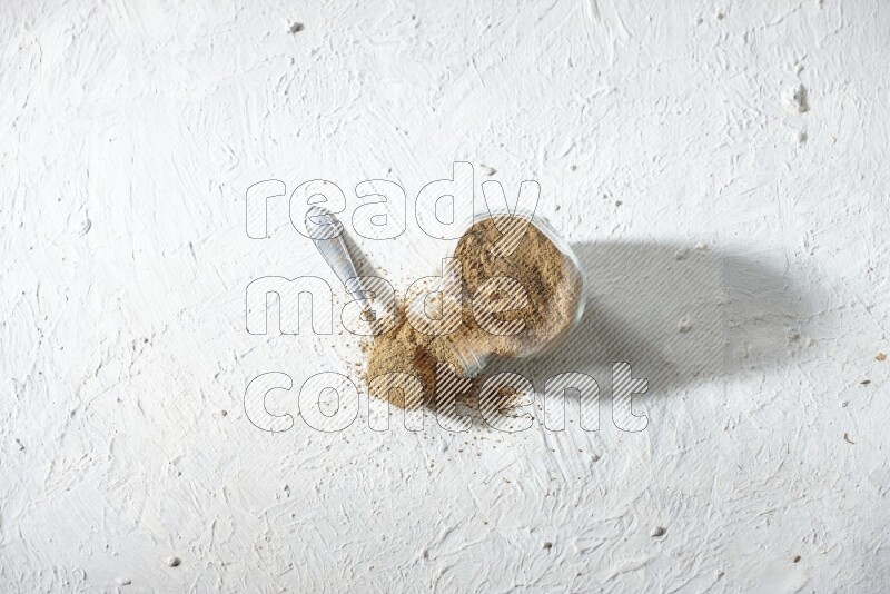 A flipped glass spice jar and a metal spoon full of cumin powder and powder spilled out on textured white flooring