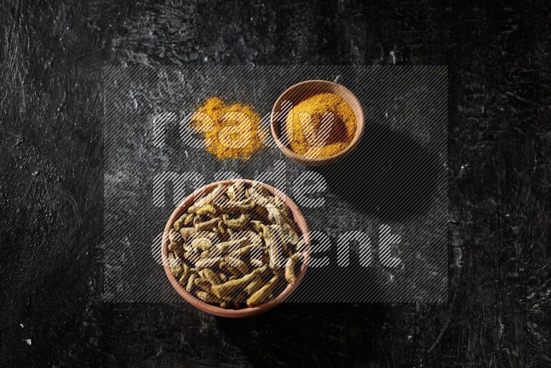 2 wooden bowls, one full of turmeric powder and the other full of dried turmeric whole fingers on a textured black flooring
