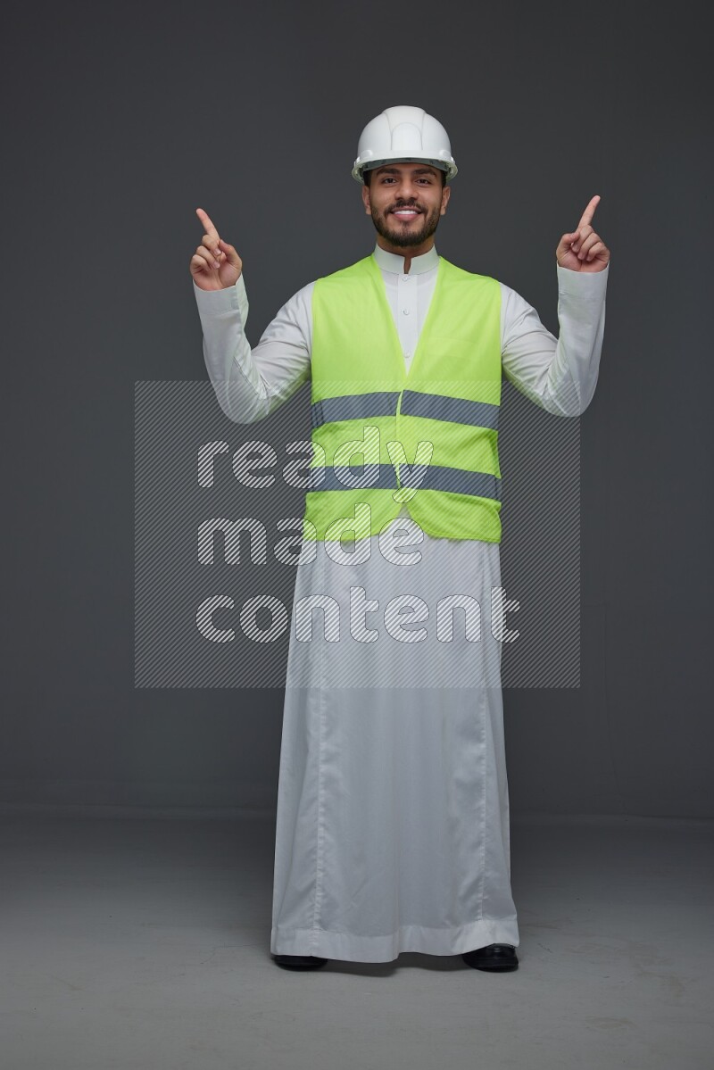 A Saudi man wearing Thobe with a yellow safety vest and white helmet standing and pointing different angles eye level on a gray background
