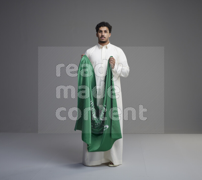 A Saudi man standing wearing thob holding big Saudi flag on gray background