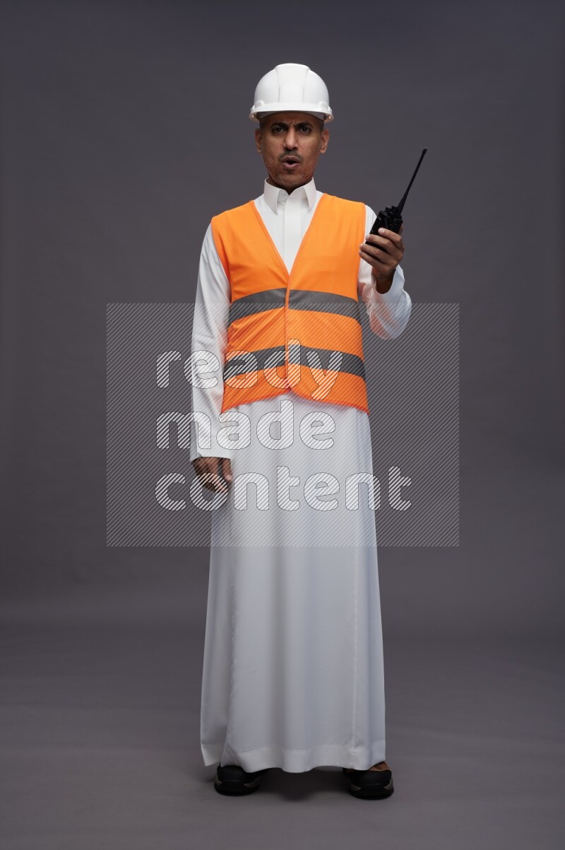 Saudi man wearing thob with engineer vest standing holding walkie-talkie on gray background