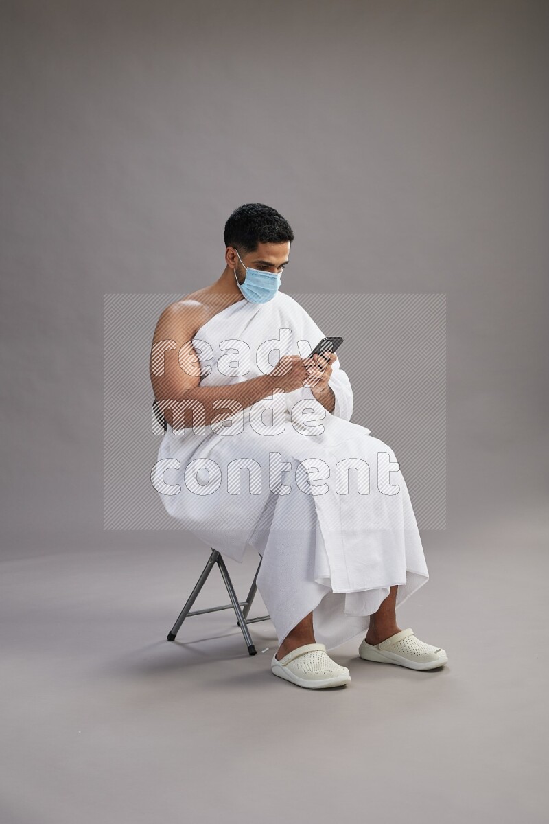 A man wearing Ehram with face mask sitting on chair texting on phone on gray background