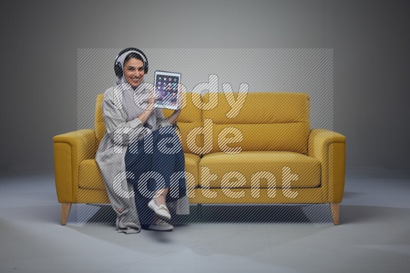 A Saudi female wearing a light gray Abaya and head scarf sitting on a yellow sofa and showing her tablet's screen while wearing headphones eye level on a grey background
