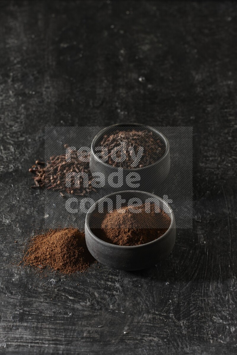 وعاءين من الفخار الأسود أحدهما ممتلئي بحبوب القرنفل والآخر ممتلئ ببودرة القرنفل على خلفية سوداء