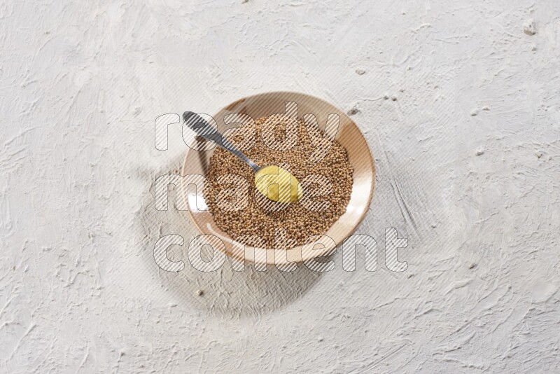 A beige pottery plate filled with mustard seeds with a metal spoon full of mustard paste in it on a textured white flooring