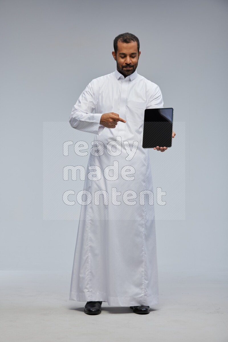 Saudi man wearing Thob standing showing tablet to camera on Gray background