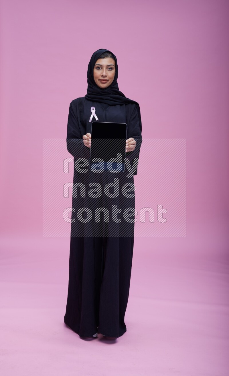 Saudi woman wearing pink ribbon on Abaya standing showing a tablet to camera on pink background