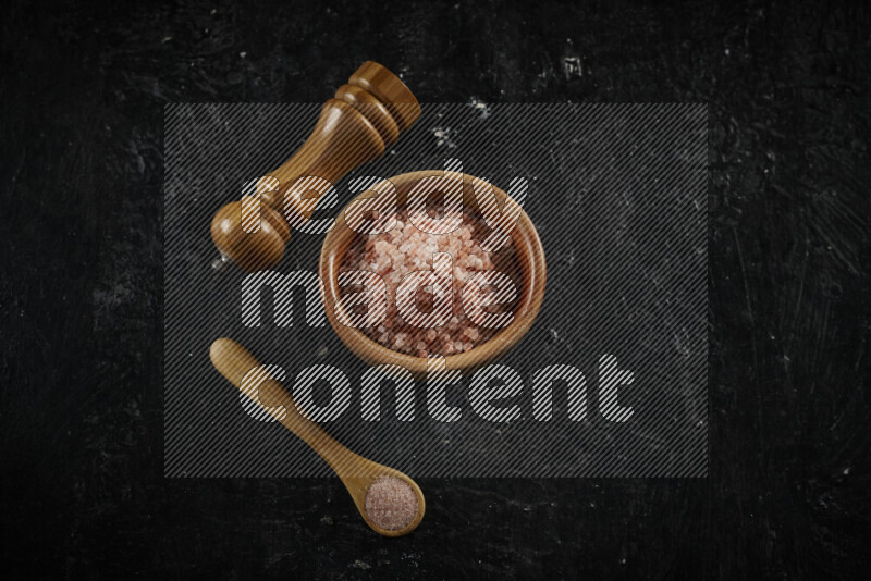 A wooden bowl and spoon filled with coarse pink himalayan salt and a wooden grinder beside them on black background