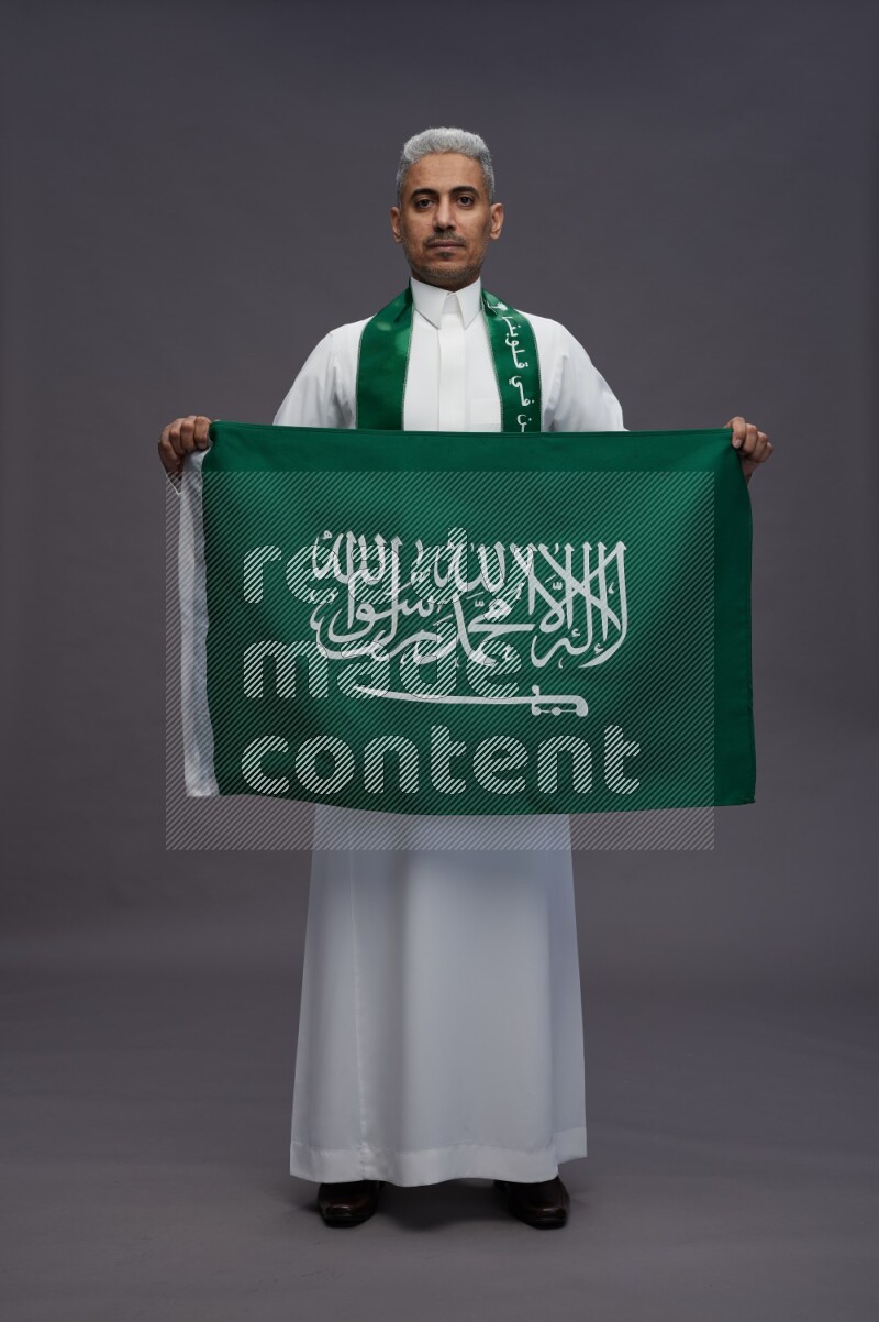 Saudi man wearing thob standing holding Saudi flag on gray background