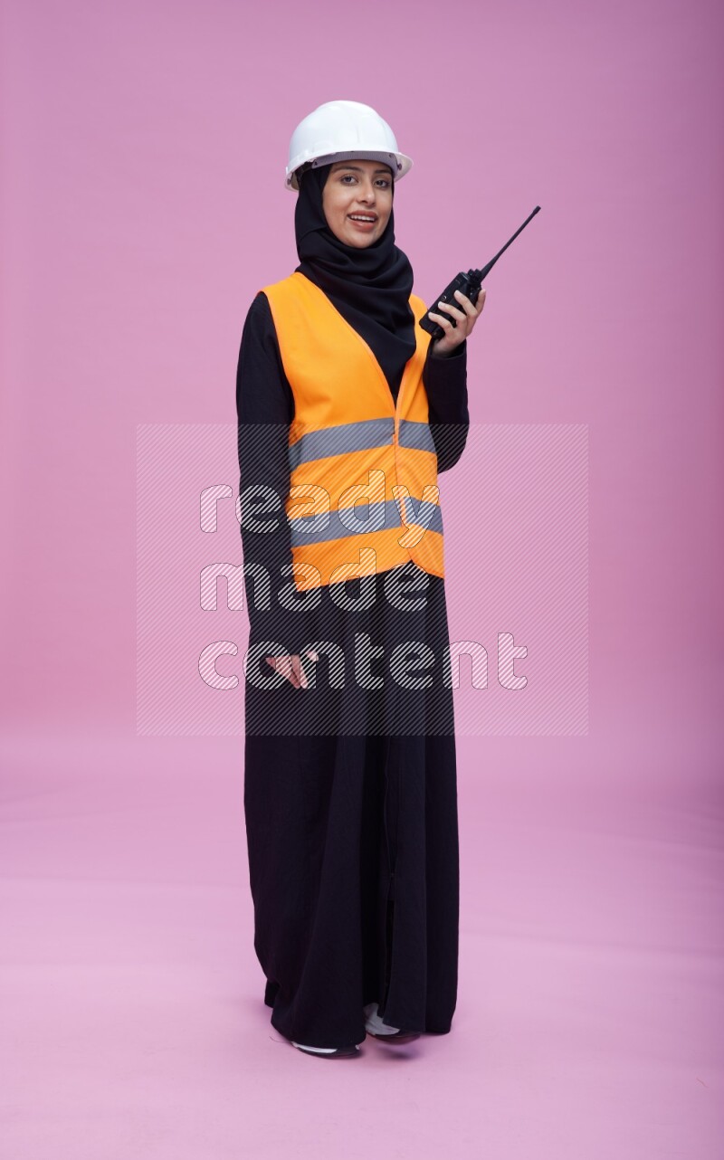 Saudi woman wearing Abaya with engineer vest and helmet standing holding walkie-talkie on pink background