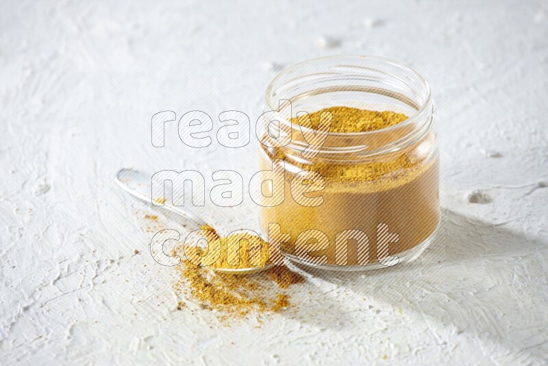 A glass jar and a metal spoon full of turmeric powder on a textured white flooring